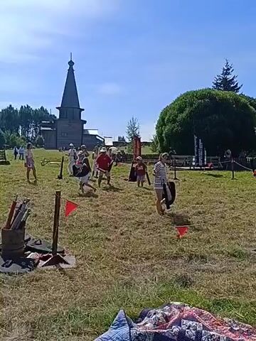 A sunny outdoor daytime scene features a wide grassy field with a tall wooden building resembling a church or tower in the background to the left, and a large bushy tree to the right. Several individuals are spread across the field, many holding what appear to be wooden swords and shields. In the foreground to the left, a metal bucket filled with poles or sticks stands on the grass. Red flags on stands are visible at various points on the field. Some people appear to be engaged in simulated combat or practicing with the props, while others observe or move about the area. The sky is clear and blue.