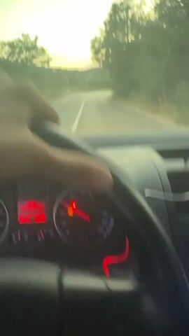 The video begins with a first-person perspective inside a car, focusing on a hand gripping the steering wheel. The dashboard instruments are visible, with a red needle indicating speed. Outside the windshield, a road curves ahead, surrounded by trees and vegetation. The sky displays a soft sunset with hues of orange and yellow. The camera perspective shifts slightly, revealing more of the dashboard and a suction-cup mount for a device on the windshield. Dust or small particles are visible on the windshield. The car appears to be driving along a rural road as the sun begins to set.