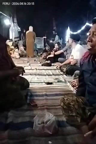 A group of people are seated on the ground on mats at night, under what appears to be a tent or canopy. Several individuals are smoking cigarettes, with visible plumes of smoke. A timestamp in the upper left corner reads "FERU | 2026.08.16 20:13". The people are casually dressed. In the foreground, there is a plastic bag filled with what looks like cigarette butts or other trash. The lighting is dim, suggesting artificial lighting from above. The atmosphere appears relaxed, with people engaged in conversation or simply sitting together.
