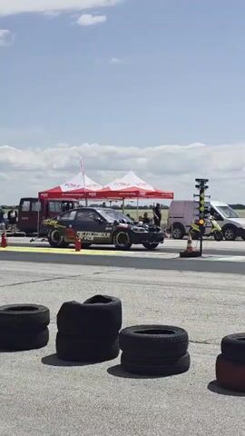 The video shows a daytime outdoor scene at a race track. In the foreground, there are stacks of tires on the asphalt. In the middle ground, a dark-colored performance car, adorned with graphics and sponsor logos, is positioned on the track, seemingly preparing for a race. Several people are standing near the car, some under a tent. In the background, more people are visible, and beyond them, fields and hills under a cloudy blue sky. The car then accelerates down the track. Another dark-colored car is seen in the far distance further down the track. The perspective shifts throughout the video, showing the race car from different angles as it moves away from the camera.