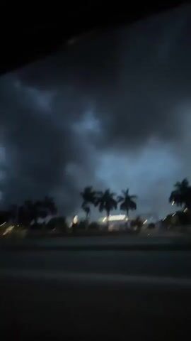 The video shows a night scene from inside a moving vehicle, likely a car. The sky is dominated by thick, dark storm clouds, with hints of blue showing through in some areas. Below the clouds, palm trees are visible in silhouette along the roadside. There are lights lining the road, appearing blurry due to the movement of the vehicle. The overall atmosphere is dark and ominous, suggestive of an impending storm.
