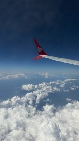 This is a point-of-view video taken from an airplane window, looking out at the sky and clouds. The right wing of the airplane is visible in the foreground, with a red tip that features a logo. The sky is a deep blue, transitioning to a lighter blue near the horizon, and is dotted with white, puffy clouds. Below the airplane, a vast expanse of clouds stretches out, with glimpses of what appears to be land or sea visible through gaps in the cloud cover. The overall impression is of flying at a high altitude on a clear day. There are no people or specific actions depicted beyond the context of being in an airplane.