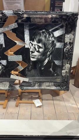 The video shows a piece of art, likely a mixed media painting or collage, leaning on two wooden sawhorse stands. The artwork has a black and white image of the two members of the band Oasis, likely Liam and Noel Gallagher, over a Union Jack flag. A zigzagging shape composed of reflective metal and brown material cuts through the image. The frame of the artwork is ornately decorated in black. A "Certificate of Authenticity" is visible behind the artwork. The surface underneath the artwork is a pale wooden floor. The shot appears to be taken indoors, possibly in a gallery or studio.