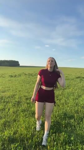 A woman with long, light-brown hair is walking through a grassy field on a sunny day. She is wearing a maroon athletic set consisting of a short-sleeved top and shorts, along with white socks and athletic shoes. The field is green with occasional yellow flowers, and a line of trees is visible in the distance. The sky is blue with scattered clouds. The camera follows her as she walks, alternating between showing her face and her back. At one point, she looks up at the sky.