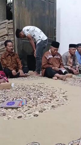 The video shows four men sitting on the floor facing a rug with ornate patterns. In the background, a younger man stands bending over. The seated men are wearing traditional clothing, including peci hats. One of the seated men is reading a book. The environment appears to be indoors, with wooden doors and walls visible.