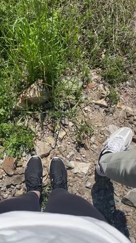 The video shows a scenic view of a mountainous landscape. It begins with a ground-level shot of two pairs of feet on a rocky, grassy terrain. One pair of shoes is black and the other is white with khaki pants. The camera then pans upwards to reveal a lush green valley with rolling hills covered in trees. The sky is clear and blue. The overall impression is of a beautiful, natural environment.
