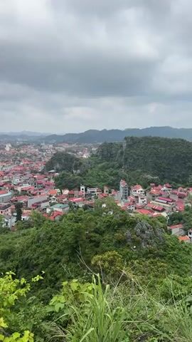 A panoramic view of a densely populated town is captured from a high vantage point. Numerous red-roofed houses are clustered together, interspersed with taller modern buildings. Lush green foliage, including trees and bushes, covers the foreground and climbs up the rocky hillsides surrounding the town. In the distance, a hazy landscape of rolling hills and mountains stretches to the horizon under a cloudy sky. A body of water, possibly a lake or river, is visible within the town. The scene is static, with no visible people or actions.