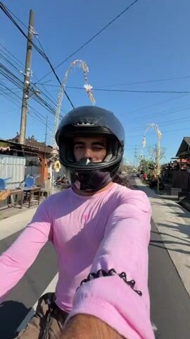 A man wearing a black helmet and a pink long-sleeved shirt rides a motorcycle down a paved street. He is looking at the camera, which appears to be mounted on the motorcycle, and smiling. The street is lined with buildings and trees, and there are power lines overhead. The sky is clear and blue.