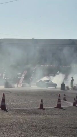 A car is drifting on a paved surface, producing a large amount of smoke. The car is dark-colored and appears to be a sedan or coupe. The drifting event is taking place outdoors during the daytime, with a clear sky and sunlight visible. The ground is covered in asphalt or gravel. Orange and white traffic cones are placed around the course to mark the path or create obstacles. In the background, there are spectators and some temporary structures or banners, possibly associated with a racing or drifting event. The overall scene suggests an organized automotive event focused on drifting.