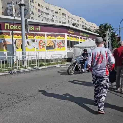 In an outdoor urban setting, two individuals are seen riding motorcycles. The first motorcycle, a cruiser with saddlebags, is ridden by a person wearing a helmet, sunglasses, a dark vest over a grey shirt, and blue jeans. The second motorcycle, a dark sport-touring bike with saddlebags and a top case, is ridden by a person wearing a black helmet with a tinted visor, a dark vest over a white t-shirt, and grey jeans. They are proceeding on an asphalt road with a sidewalk and a white metal fence to their left. Behind the fence is a building with a sign that reads "Пекарня №1" (Bakery No. 1) and features images of baked goods and pizzas. In the background, a multi-story building with many windows is visible, along with trees and a white tent structure. The weather appears to be sunny, casting shadows on the road. To the right of the motorcycles, two other individuals are partially visible, one in a red shirt and another person walking away from the camera wearing a patterned shirt and camouflage pants. The scene suggests a daytime event or gathering, possibly a motorcycle rally or a street fair.