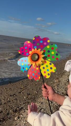 A person holds a colorful pinwheel with a sunflower design in the center. The pinwheel has heart-shaped petals adorned with polka dots in various bright colors. The person is on a beach, with the ocean and a sandy shore visible in the background. The sky is blue with some clouds. A baby is partially visible in the foreground, wearing a white hat and a light-colored sweater. The baby's hands are extended, possibly reaching for the pinwheel or the water. The pinwheel is spinning in the wind.
