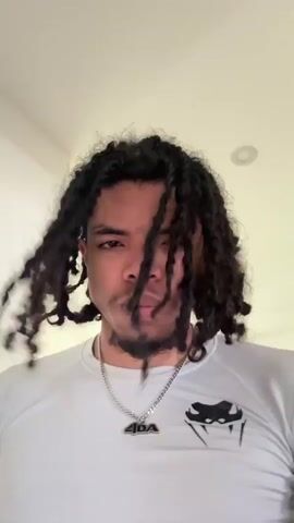 This is a short video featuring a young man with dark, curly dreadlocks. He is wearing a light gray t-shirt with a black logo on the left side of his chest. Around his neck, he wears a silver chain with a pendant. The background is slightly blurred, but appears to be an indoor setting with light-colored walls and doorways, suggesting a home or apartment. The lighting is bright, indicating daytime. The man's expression is neutral, with a slight smirk. There are no other people or objects of note visible in the frame. The video appears to be a selfie or a personal recording, possibly for social media. No specific activity is taking place other than the man looking at the camera.