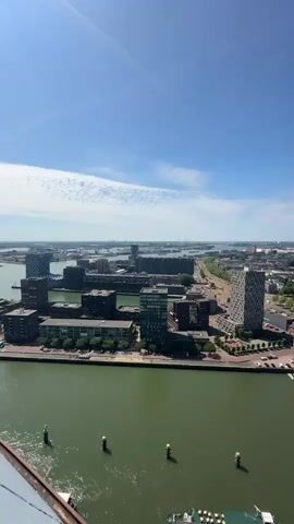 This is a vertical video taken from a high vantage point overlooking a city and a body of water. The sky is clear and blue with a few wispy white clouds. In the foreground, the greenish water of a harbor or river is visible, with several yellow buoys. Part of a structure, possibly a railing or the edge of a building, is visible in the bottom left corner of the frame. Across the water, a cityscape unfolds with numerous buildings of varying heights and architectural styles. Some buildings are modern, featuring dark facades and geometric designs, while others appear older with brick exteriors. A wide road or pathway runs parallel to the waterfront, with trees lining parts of it. Further in the distance, more buildings and industrial structures can be seen, extending towards the horizon. Several boats are docked along the waterfront. The overall scene depicts an urban environment with a significant body of water, suggesting a coastal or riverside city.