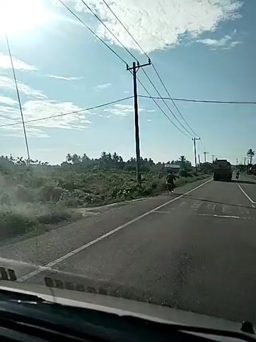 The video shows a view from inside a vehicle, looking out at a road. A person on a bicycle is riding on the side of the road, to the left of the vehicle. There is a pedestrian crossing marked on the road. Utility poles and power lines are visible along the side of the road. A truck is driving in the distance on the road, moving away from the camera. The environment appears to be rural, with vegetation and some buildings in the background. The sky is blue with some clouds.