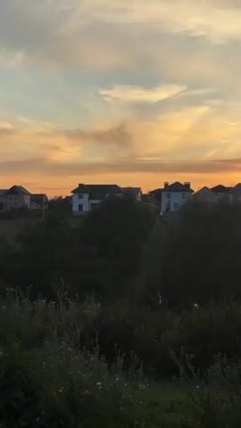 A time lapse video shows a sunset over a small village with houses and trees. The sky is filled with colorful clouds in shades of orange, yellow, and blue. The sun is setting behind the houses, casting a warm glow over the scene. There is a small body of water in the foreground, reflecting the colors of the sky. The video is shot from a slightly elevated position, looking down on the village.