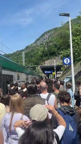 The video shows a large crowd of people standing at a train station on a sunny day. The people are waiting to board a green and silver train on the left side of the frame. The platform is crowded and there is a sign indicating "Platform 2" next to a lamp post. In the background, a mountain covered with trees is visible. The sky is clear and blue. The camera appears to be held at head height.