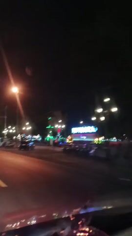 The video begins with a street-level view at night, looking towards a brightly lit sign that reads "KUTOARJO". The sign is illuminated with neon lights in blue and purple, with smaller text below it that is difficult to read but appears to be "Purworejo". There are streetlights visible, casting a warm glow on the wet asphalt road. Several cars are on the road, some appearing to be moving and others parked. A prominent streetlamp is in the foreground on the left side of the screen, with a bright lens flare. In the distance, there are more lights and the silhouette of a structure that might be a tower or monument, illuminated in green. Pedestrians are visible near the sign, and there are trees and other vegetation along the sides of the road. The camera appears to be inside a vehicle, possibly looking out the windshield or a side window, as the lower part of the vehicle's interior is visible, including what looks like the dashboard. The overall atmosphere is that of a city street at night, bustling with activity and illuminated by artificial lights. The video is shaky, suggesting it was recorded while in motion.