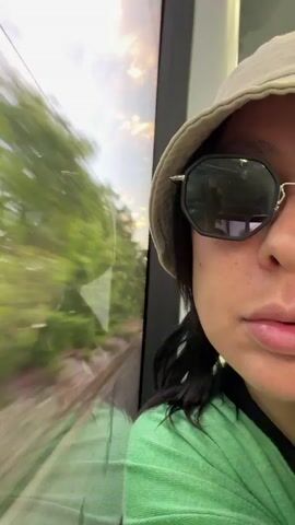 A close-up selfie shot of a woman on a train. The woman is wearing a light-colored bucket hat, sunglasses, and a bright green sweater. The train is moving, as evidenced by the blurred scenery visible through the window behind her, which includes trees, sky, and possibly a bridge or overpass. The woman looks directly at the camera for most of the video, and briefly smirks towards the end. The lighting suggests it is daytime.