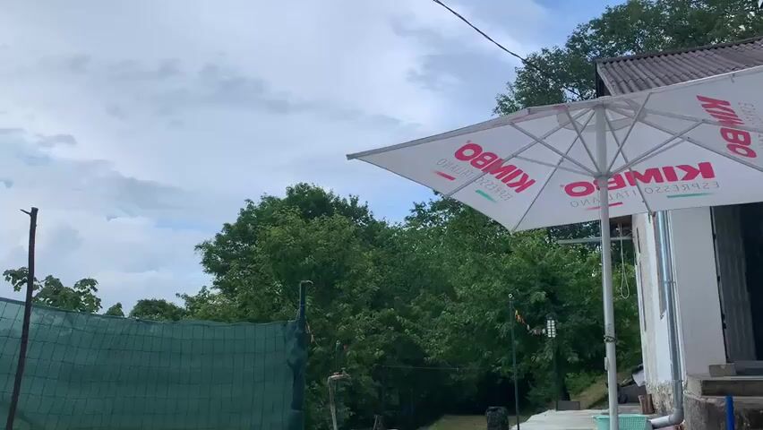 The media file shows an outdoor scene with a focus on a large, white patio umbrella with the "KIMBO ESPRESSO ITALIANO" logo printed in red. The umbrella is tilted, suggesting it might be in use or preparing for rain. In the background, lush green trees create a dense foliage. Above, the sky is filled with dramatic, heavy clouds, indicating overcast or potentially stormy weather. The lighting suggests it's daytime, but the cloud cover is obscuring direct sunlight.

Partially visible in the foreground is a green mesh fence, possibly part of a sports court or boundary. There are also some dark vertical posts, one appearing to be a light pole or similar fixture, and another that could be a support for the fence or another structure. The architecture of a building is visible on the right, with a sloping roof and stairs leading up to an entrance.

The overall impression is a slightly moody, overcast day, possibly before or after a rain shower. The scene appears to be a casual outdoor area, perhaps a backyard, patio, or a recreational space. The prominent "KIMBO ESPRESSO ITALIANO" text on the umbrella clearly identifies a brand, hinting at a commercial setting or a sponsorship. The location cues, combined with the umbrella and building style, suggest a European setting. No people are visible in the frame, leaving the focus on the environment and the branded object.