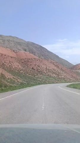 The video is a first-person perspective shot from inside a vehicle traveling on a paved road that curves to the right. The road is flanked by a gently sloping grassy embankment on the left and a rocky, arid mountainside on the right. The mountainside is characterized by layers of reddish-orange and gray soil, with sparse vegetation. The sky is clear and blue with a few wispy clouds. In the distance, on the right side of the road, a dark-colored sedan is visible, traveling in the same direction. The vehicle's interior, specifically the dashboard, is partially visible at the bottom of the frame. The overall impression is of a scenic drive through a mountainous, desert-like landscape.