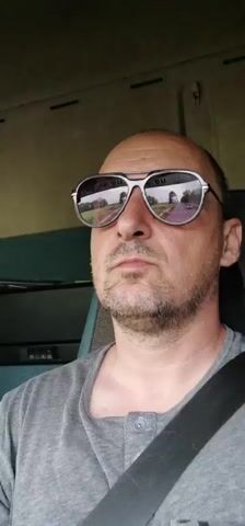A man with a shaved head and sunglasses is recording a selfie video in the driver's seat of a vehicle. He is wearing a gray t-shirt and a seatbelt. His expression is neutral. The background is slightly blurred, showing the interior of the vehicle and hints of the landscape outside. The audio contains dialogue in a language other than English.