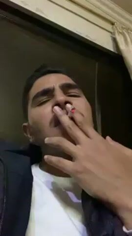 A young man with short dark hair and a mustache is seen from a low angle indoors. He wears a black puffy jacket over a white t-shirt. Initially, he holds a lit cigarette to his lips. He then moves the cigarette away and blows a plume of smoke upward. The background includes a dark surface, a patterned molding, and a light-colored curtain.