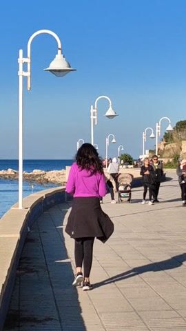 The video shows a paved walkway on a sunny day.  Along the walkway are evenly spaced lampposts. The bright blue sea is visible on the left, along with a rocky shoreline. There are multiple people on the walkway, some standing and some walking. A woman with long, dark hair and wearing a bright purple jacket, black skirt, and leggings walks in the foreground, facing the sea.  Her shadow stretches out onto the walkway.  Other people are further down the walkway, including a woman pushing a stroller. The sky is clear and bright blue. The camera is positioned so that the scene appears rotated counterclockwise.