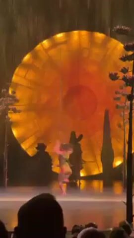A female aerial acrobat performs on a suspended, round trapeze during a stage performance. She wears a brightly colored leotard. Behind her is a large, circular backdrop illuminated in warm orange and yellow tones. Artificial rain falls on the stage around her. Trees flank the stage. A beam of green light illuminates the performer from above. The audience is partially visible in the foreground.