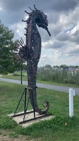 A metal sculpture of a seahorse stands tall on a grassy area next to a paved path. The seahorse sculpture is made of various metal pieces, including gears and plates, and has a dark, rusty patina. It is supported by a black metal stand on a concrete base. To the left of the sculpture, a tree with green leaves is visible. Behind the seahorse, tall reeds or grass grow, and in the distance, houses and trees can be seen under a cloudy sky. A white post stands to the right of the sculpture. The video pans slightly to the right, showing the full seahorse sculpture.
