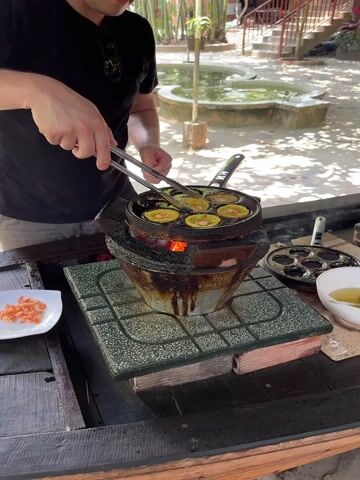 A man and a woman are cooking outdoors in Tân Thạch, Vietnam. The man, dressed in a black t-shirt and light pants, is using tongs to turn small, circular, yellow food items, possibly Vietnamese Bánh Khọt, in a cast iron pan over a charcoal stove. A small plate of chopped red shrimp is visible on the wooden table next to him.

The woman, wearing a black t-shirt with a "Simply Love" design and a black apron, is standing at an adjacent cooking station. She is pouring liquid from a white bowl into another cast iron pan on her own charcoal stove. Both individuals appear focused on their cooking tasks, with the woman showing a slight smile in one instance.

The cooking takes place on a wooden table under a shaded outdoor structure. In the background, there's a paved area with a water feature or pond, lush greenery including bamboo, and rustic buildings with stairs and arched windows, typical of a resort or traditional Vietnamese setting. The scene is brightly lit by natural sunlight, indicating it's daytime.