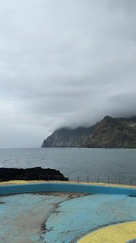 The video shows a cloudy day at the ocean. In the foreground, there is an empty swimming pool with blue and yellow paint. Beyond the pool is a rocky shoreline and then the ocean. In the distance is a large, green, mountainous landmass partially covered in clouds. The sky is overcast.