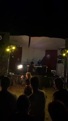 The video shows a nighttime outdoor music performance. A stage is set up with two people on it, one appearing to be playing a keyboard or synthesizer and the other standing nearby. There are lights on the stage and around the audience. A crowd of people is gathered in front of the stage, silhouetted against the lights. Some members of the audience are holding up lights. One person in the audience appears to be holding a flaming prop. The stage has a backdrop that appears to be a white cloth with some red fabric behind it. There are speakers on the stage and in front of it. The ground in front of the stage is covered with grass. The overall atmosphere is that of an outdoor concert or festival.