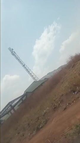The video shows a rural scene, seemingly filmed from a moving vehicle on a dirt road. The camera angle is often tilted. Initially, a tall metal structure, possibly a crane, is visible against a sky with scattered clouds. The foreground consists of a reddish-brown, rocky terrain. A man rides a motorbike on the road. A large truck with a covered bed passes by. Further along, the video reveals a row of buildings with corrugated metal roofs, situated on the slope of a hill. The architecture appears simple and functional. Vegetation, consisting of trees and dry grass, is sparse. The overall impression is of a somewhat arid, undeveloped area.