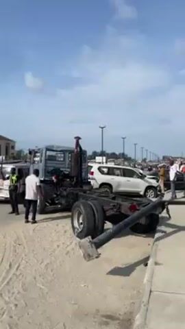 The video shows a street scene in what appears to be an urban area, possibly near a beach, indicated by the sand near the road. Several vehicles are visible, including a truck frame missing its body, a white SUV, and a white van. People are present, some appearing to be interacting near the vehicles, including one individual wearing a green uniform that suggests they may be a traffic officer. A fallen light pole lies in the foreground. The sky is blue with scattered clouds.