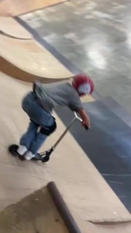 A young person on a scooter, wearing a red helmet, blue jeans, and knee pads, is performing a trick in a skate park. The person rides up a ramp and then launches into the air, performing a flip with the scooter. The skate park has wooden ramps and walls, with metal and black rubber surfaces on some of the ramps. Fluorescent lights are visible on the ceiling.
