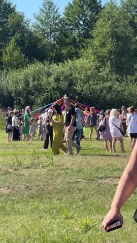 The video shows a sunny outdoor gathering with a group of people around a maypole decorated with colorful ribbons. In the foreground, a woman with her hair in a bun is visible from the side, wearing a black strapless top and dark pants. The background is filled with trees and lush green grass. People of various ages are seen milling around the maypole, some appearing to be dancing or interacting. The overall atmosphere seems festive and relaxed, with a bright blue sky overhead.