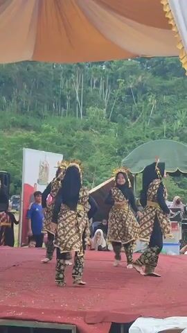 A group of young females, dressed in traditional patterned attire and headscarves, performs a dance on a red stage. The stage is covered with a red carpet, and a large tent with orange and white fabric is visible overhead, providing shade. In the background, a lush green hillside is covered with trees. Several spectators are present, some are partially visible behind the dancers. The dancers are moving in unison, with some raising their arms and others bending their knees, indicating a coordinated performance. The lighting suggests it is daytime, likely outdoors.