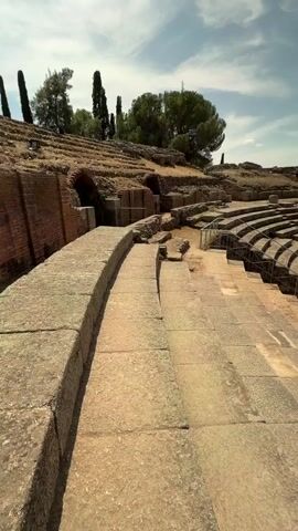 The video shows a panoramic view of an ancient Roman amphitheater on a sunny day. The camera pans from a close-up of stone seating in the foreground to reveal the full structure of the amphitheater, including the stage area and tiered seating. The stonework is weathered and shows signs of age. Beyond the amphitheater, trees and a small town with buildings are visible under a clear blue sky with some scattered white clouds. A few people are walking around the amphitheater, appearing to be tourists. The scene is bathed in bright sunlight, casting shadows on the stone structures.
