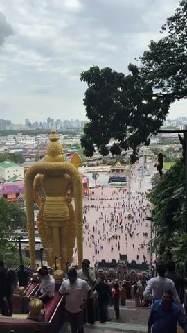 The video shows a view of the Batu Caves in Malaysia. The camera is positioned on the steps leading up to the caves, facing away from the cave entrance. In the foreground, there's a large golden statue of the Hindu deity Murugan. Many people are seen walking up and down the colorful stairs. The steps have red, pink, green, and blue paint on them. The background features a cityscape with numerous buildings, trees and foliage. There is a man in a red shirt who is looking at his phone. Overall, the scene captures the vibrant and busy atmosphere of the Batu Caves, with its mix of religious significance and tourist attraction.