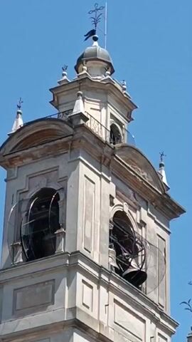 The video shows a church bell tower with two large bells visible in arched openings. The tower is made of light-colored stone and has a classical design with moldings and details. At the top is a small dome-shaped structure with a weather vane. The sky is blue. The bells are ringing.