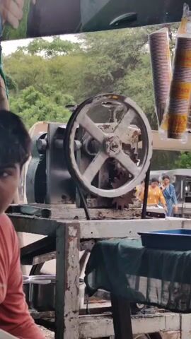 A young person wearing a pink shirt sits in front of a sugarcane juice machine. The machine has a large white wheel that is spinning, and a metal crank on top. Sugarcane is visible next to the machine. There are paper cups stacked on the right side of the frame. In the background, there are trees and a few people. The environment appears to be outdoors, possibly a street vendor stall.