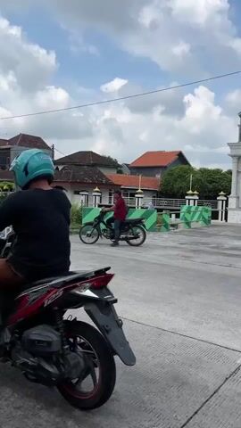 The video shows a red motorcycle with a rider wearing a blue hoodie and a white helmet. The motorcycle is on a paved surface, possibly a road or a parking lot, with a building in the background. Another person on a bicycle is visible in the distance. The sky is cloudy. The scene appears to be outdoors, possibly in a village or town.