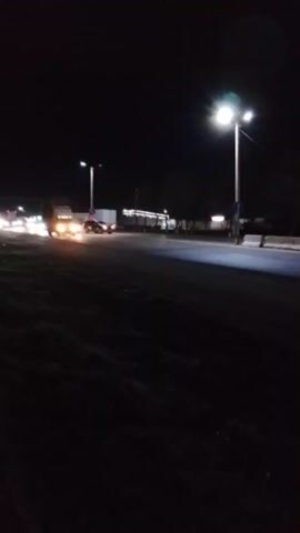 The video captures a nighttime street scene, viewed from a low, slightly angled perspective. The darkness of the night is broken by artificial illumination from multiple streetlights lining the road on the left side of the frame. These streetlights cast bright pools of light onto the wet or reflective asphalt road surface.

In the distance, further down the road on the left, a cluster of lights suggests the presence of buildings, possibly commercial establishments or a gas station. Various vehicles are visible on the road. Initially, a car with bright headlights is seen driving away from the camera. Later, a dark-colored car with its headlights on approaches and passes the camera, its taillights becoming visible as it continues down the road. Other car headlights and taillights can be seen further in the distance, indicating ongoing traffic.

To the right of the road, a dark, grassy bank or shoulder is visible, with some indistinct foliage. The overall atmosphere is one of an active road at night. The audio features the distinct sound of a car engine and tires as a vehicle passes by. The video appears to have been recorded vertically, giving the impression of the scene being rotated 90 degrees clockwise.