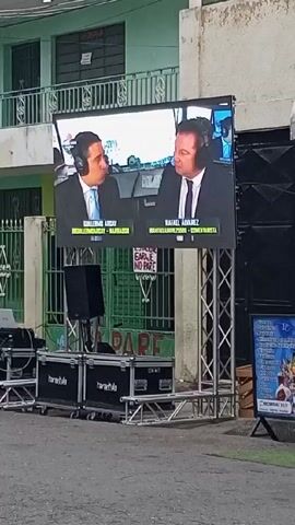 The video shows a street scene with people gathered on a sidewalk and a large screen displaying a news or commentary program.

The screen is set up on a metal frame with sound equipment below. It displays two men in suits, Guillermo Arcay and Rafael Alvarez, likely news anchors or commentators. The audio indicates they are having a conversation, mentioning work and giving arms a rest.

On the sidewalk, a group of people are sitting and standing, some talking to each other. A woman in a blue shirt appears to be interacting with them.

The street has a car and a motorbike passing by. There are buildings on both sides of the street, and the overall atmosphere seems like a community gathering to watch the broadcast.