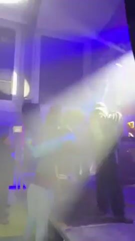 The video shows a group of people standing and moving around in a dark, blue-lit space. There's a sound system with speakers visible. The people are partially obscured by the low lighting and are not clearly identifiable. A bar area or table setup can be inferred based on the visible elements. The setting appears to be a club or entertainment venue with a sound system.