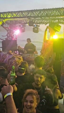 The video shows a music event taking place near a beach. Initially, a crowd of people is visible in front of a stage with a DJ playing music. Several individuals are smoking, and the atmosphere appears lively. The camera then pans up to reveal the ocean, with waves gently crashing on the shore under a pastel-colored sky. As the camera continues to pan, a few people are seen raising their hands in the air, enjoying the view.  The scene conveys a sense of a party happening in a scenic location.