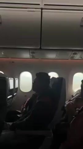 The video shows the interior of an airplane cabin. A man wearing glasses is sitting in a window seat, looking out. Other passengers are visible in the background. The plane appears to be approaching a city, visible through the windows. The cabin is dimly lit, and overhead storage compartments are visible. The scene appears calm and uneventful.