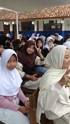 A group of young women, most wearing hijabs, are seated in chairs outdoors. They appear to be attending an event. In the foreground, three young women are visible. The woman on the left, wearing a white hijab and pink shirt, looks toward the camera with a neutral expression. The woman in the center, wearing a brown hijab, smiles and makes a peace sign with her hand. The woman on the right, wearing a light-colored hijab, also smiles and looks toward the camera. In the background, more young women are seated, some waving or looking around. Behind them, a building with a tiled roof and windows is visible. The overall atmosphere appears casual and social.