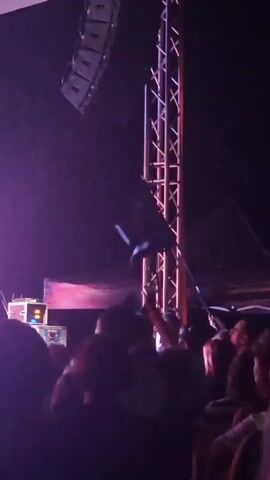 The video shows a crowd of people at what appears to be a concert or event. There are speakers mounted high on a structure, and a tall metal truss in the center of the frame. A person in the crowd is throwing a flag into the air. The flag is black with a white logo. The lighting is predominantly purple.