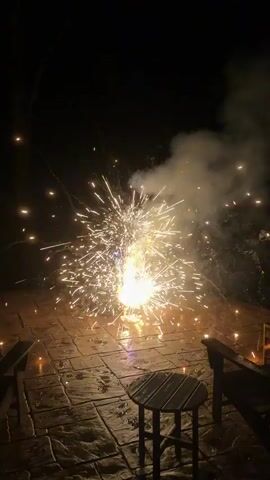 A firework erupts on a wet, stone patio at night, casting sparks and smoke into the dark sky. The patio is furnished with two wooden chairs and a small round wooden table.